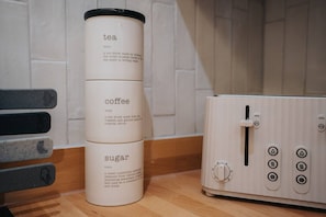 Eine Küche mit einer Kaffeemaschine, einer Kiste mit Aufgüssen und Süßungsmitteln, sowie einer Holzauskleidung.