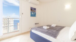 A bedroom with a large window, a bed with a blue bed skirt, a gray blanket, and a white pillow. - Beautiful Apartment Near Monaco (Beausoleil)