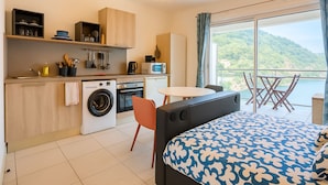 Habitació d'hotel senzilla amb cuineta, un llit amb llençols estampats, rentadora, zona de menjador amb taula i cadires, i un balcó amb vista a vegetació.