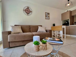 Ein Wohnzimmer mit braunem Sofa, einem runden Couchtisch mit zwei Pflanzen, einer Wanduhr und einer Küchenzeile im Hintergrund.