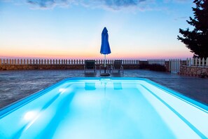 A swimming pool with a blue umbrella, stone wall, and a view of the sunset.