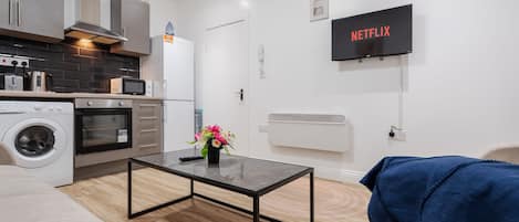 Cocina moderna con electrodomésticos, comedor con mesa, sofá y televisor mostrando 'NETFLIX'.
