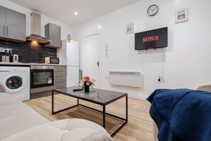 Cozinha moderna com eletrodomésticos, área de jantar com mesa, sofá e televisão exibindo 'NETFLIX'.