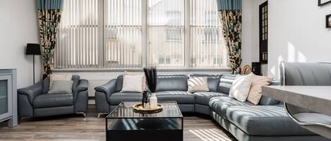 A modern living room with a sectional sofa, a glass coffee table, and a view of the outside through large windows with blinds.