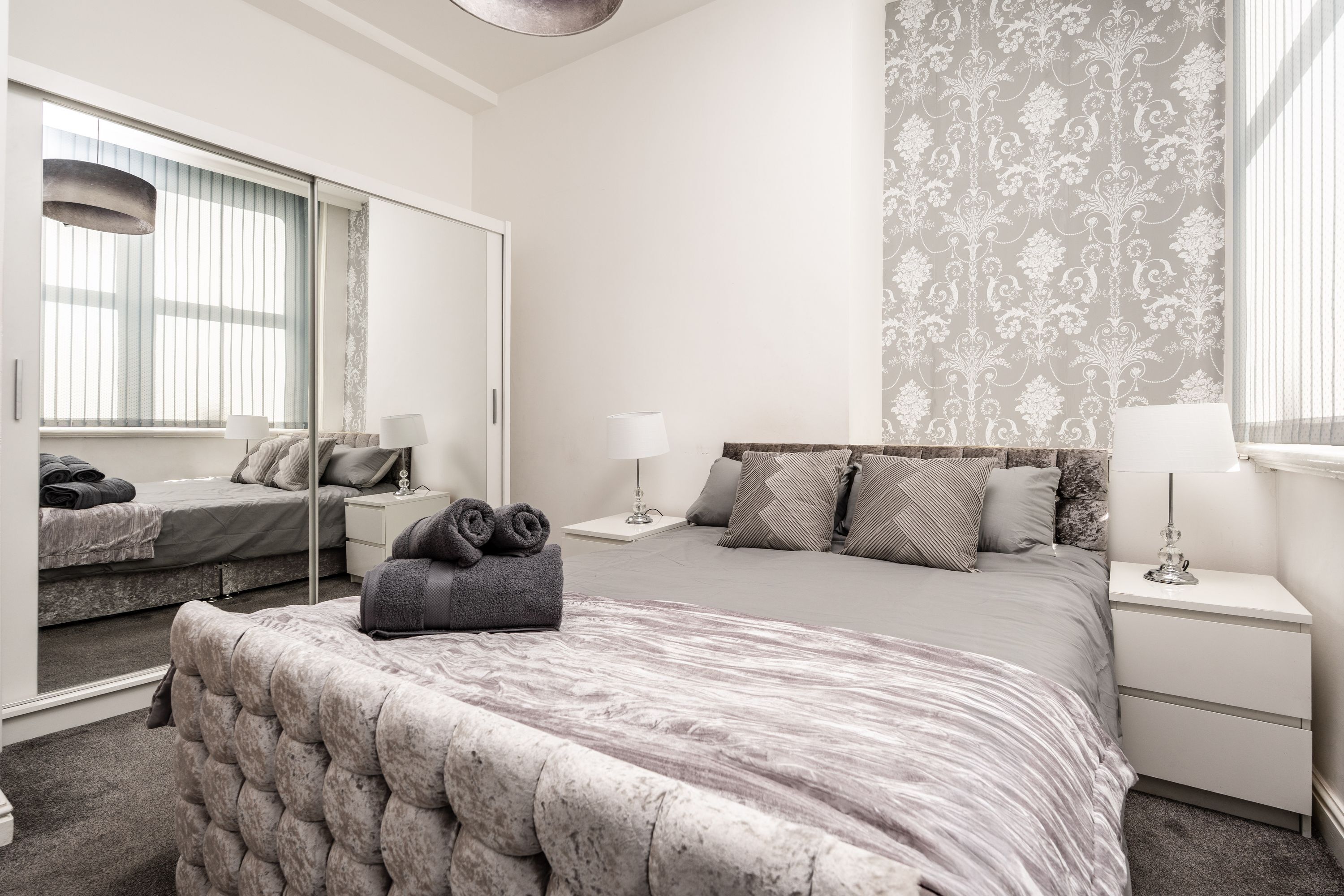 A bedroom with a large mirror, a bed with a tufted headboard, a nightstand, and a wall with a patterned wallpaper.