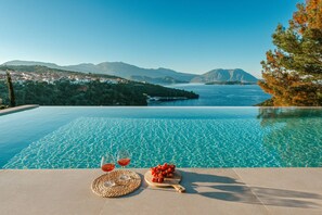 Ein Pool ohne erkennare Kante mit klarem Wasser, eine Holzplanke mit Trauben und zwei Gläsern Rotwein.