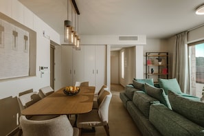 A modern living room with a wooden dining table, beige sofas, and a bookshelf. - Kohyli Villas- Villa Two With Private Pool (Meganisi)