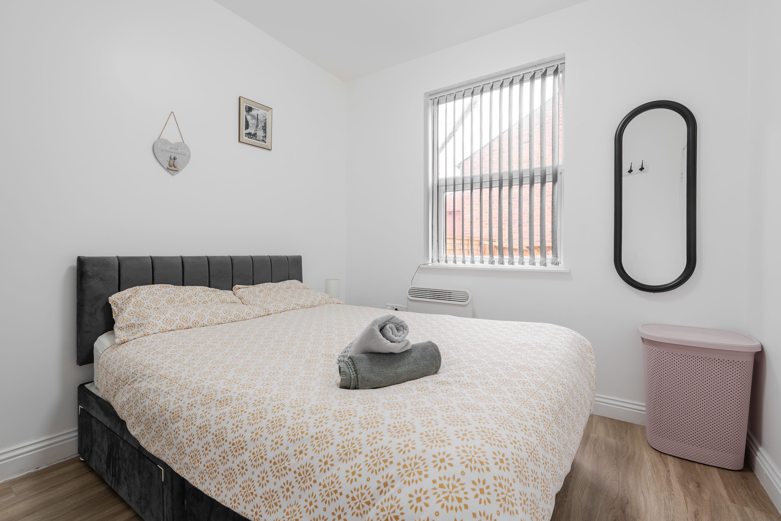 A neatly made bed with a patterned bedspread, a bedside mirror, a window with blinds, and a trash bin.