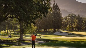 A golf course with a player in an orange shirt preparing to swing, surrounded by trees and a hilly landscape. - Luxe Napa Escape King Bed Near Top Wineries (Napa)