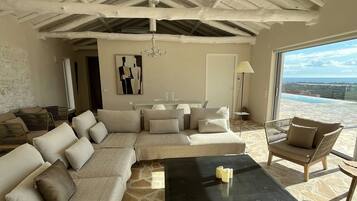 A spacious living area with a high ceiling, stone flooring, a sectional sofa, a coffee table, and a view of the ocean.