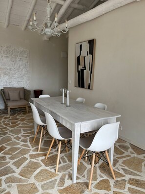 A dining area with a long table, white chairs, a chandelier, and a stone-patterned floor. - Pasparo Estate in Samos (Samos)