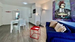 A modern living room with a blue sofa, a red side table, a dining area with white chairs, and a kitchen with white cabinets. - Charming Studio at the Gates of Monte Carlo Per (Beausoleil)