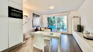 A modern kitchen with white cabinets, a dining table with chairs, a large window with a view of greenery, and a flat-screen TV on a stand. - Charming Studio at the Gates of Monte Carlo Per (Beausoleil)