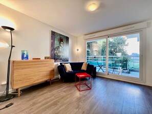 A modern living room with a wooden cabinet, a dark blue sofa, a red stool, and a large window with a view of greenery. - Charming Studio at the Gates of Monte Carlo Per (Beausoleil)