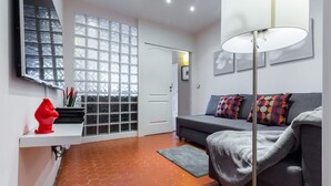 Ein modernes Wohnzimmer mit einem grauen Sofa, farbenfrohen Kissen, einer roten Skulptur, einem weißen Regal, einem großen Fenster mit Glasbausteinen und einem Boden mit sechseckigen Fliesen.