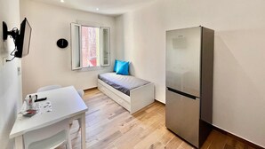 Ein kleines Zimmer mit Bett, einem kleinen Tisch, einem Kühlschrank, einem Fenster und einer Wand-Klimaanlage.