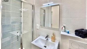 Ein modernes Badezimmer mit Duschkabine aus Glas, einem weißen Waschbecken, einem Spiegel und einem Ablagefach für Pflegeprodukte.
