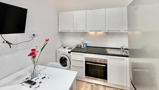 Eine kompakte Küche mit weißen Schränken, einer Waschmaschine, einem Backofen, einem Spülbecken und einem an der Wand befestigten Flachbildfernseher.
