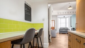 A modern kitchen with a chalkboard sign, a dining area with chairs, and a view into a living room with a sofa and a sign reading 'LIVERPOOL'. - 10 Mins to Crosby Beach Ensuite WC Parking (Liverpool)