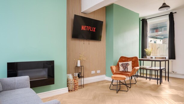 A modern living room with a flat-screen TV mounted on a wooden wall, a grey sofa, an orange armchair, and a small table with a potted plant. - 10 Mins to Crosby Beach Ensuite WC Parking (Liverpool)