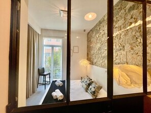 Hotellihuone, jossa on sänky, tuoli, pieni pöytä, parveke, jolta on näkymä, ja kiviseinä.