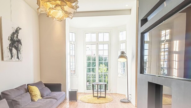 A modern living room with a grey sofa, a painting, a floor lamp, a television, and a large window with a view of greenery. - Elegant Bel Epoque Apartment in Center of Menton (Menton)