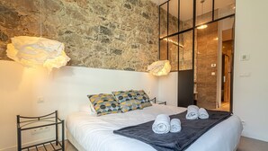 A modern bedroom with a stone wall, a bed with white bedding, patterned pillows, a black throw blanket, and rolled towels. - Elegant Bel Epoque Apartment in Center of Menton (Menton)