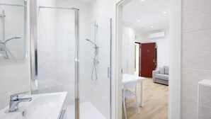 A modern bathroom with a glass-enclosed shower, a white sink, and a visible door leading to a living area with a white table and a sofa. - Cozy Studio in the Heart of Menton Walk Everywhe (Menton)