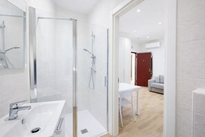 A modern bathroom with a glass-enclosed shower, a white sink, and a visible door leading to a living area with a white table and a sofa. - Cozy Studio in the Heart of Menton Walk Everywhe (Menton)