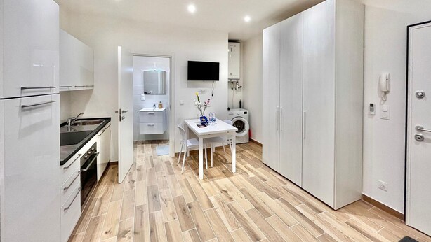 A compact kitchen with a sink, stove, and cabinets, a small dining table with chairs, a washing machine, and a TV mounted on the wall. - Cozy Studio in the Heart of Menton Walk Everywhe (Menton)