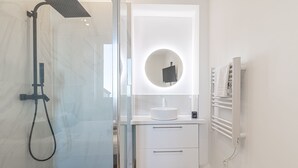 Un baño moderno con ducha compartimentada en vidrio, un tocador blanco con espejo redondo y un perchero para toallas.