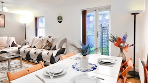 Zona pranzo con tavolo bianco imbandito per quattro persone, divano con cuscini decorati e un vaso con fiori.