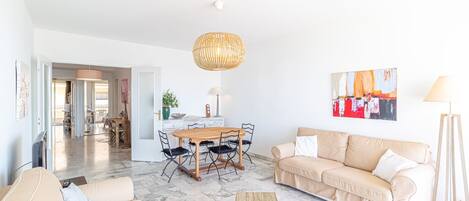 A spacious living area with a wooden dining table, beige sofas, a woven coffee table, and a hanging light fixture.