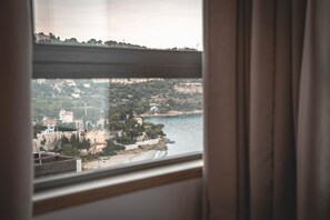 A coastal view through a window with curtains, featuring buildings, trees, and a beach. - Duplex Sea View Near Monaco AC Parking (Roquebrune-Cap-Martin)