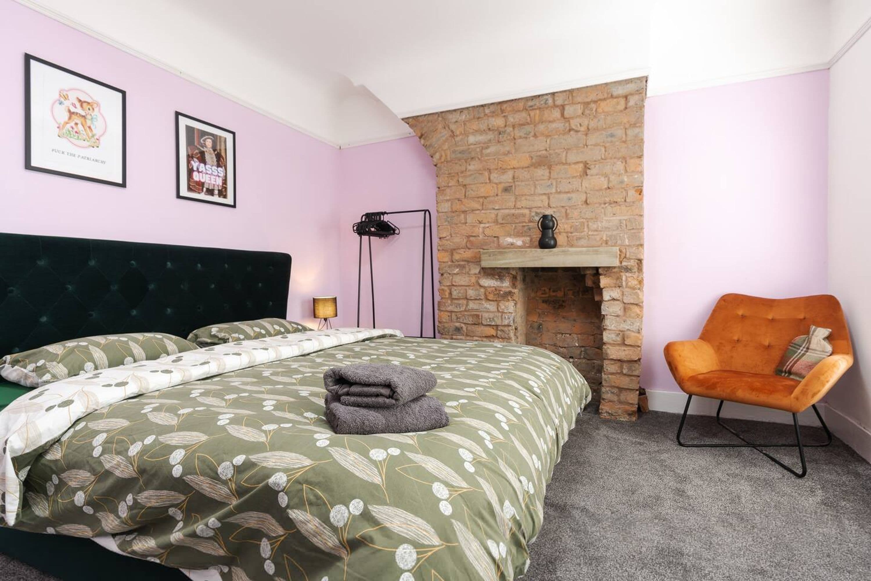 A bedroom with a bed, a brick fireplace, a chair, and two framed pictures on the wall.
