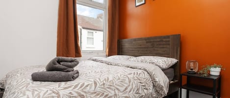 Un dormitorio con cama, mesita de noche, ventana con cortinas e imágenes enmarcadas en la pared.