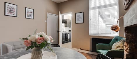 Una sala de estar con una mesa redonda, un sofá, un sillón verde y una chimenea. Hay cuadros enmarcados en la pared y una puerta que conduce a otra habitación.