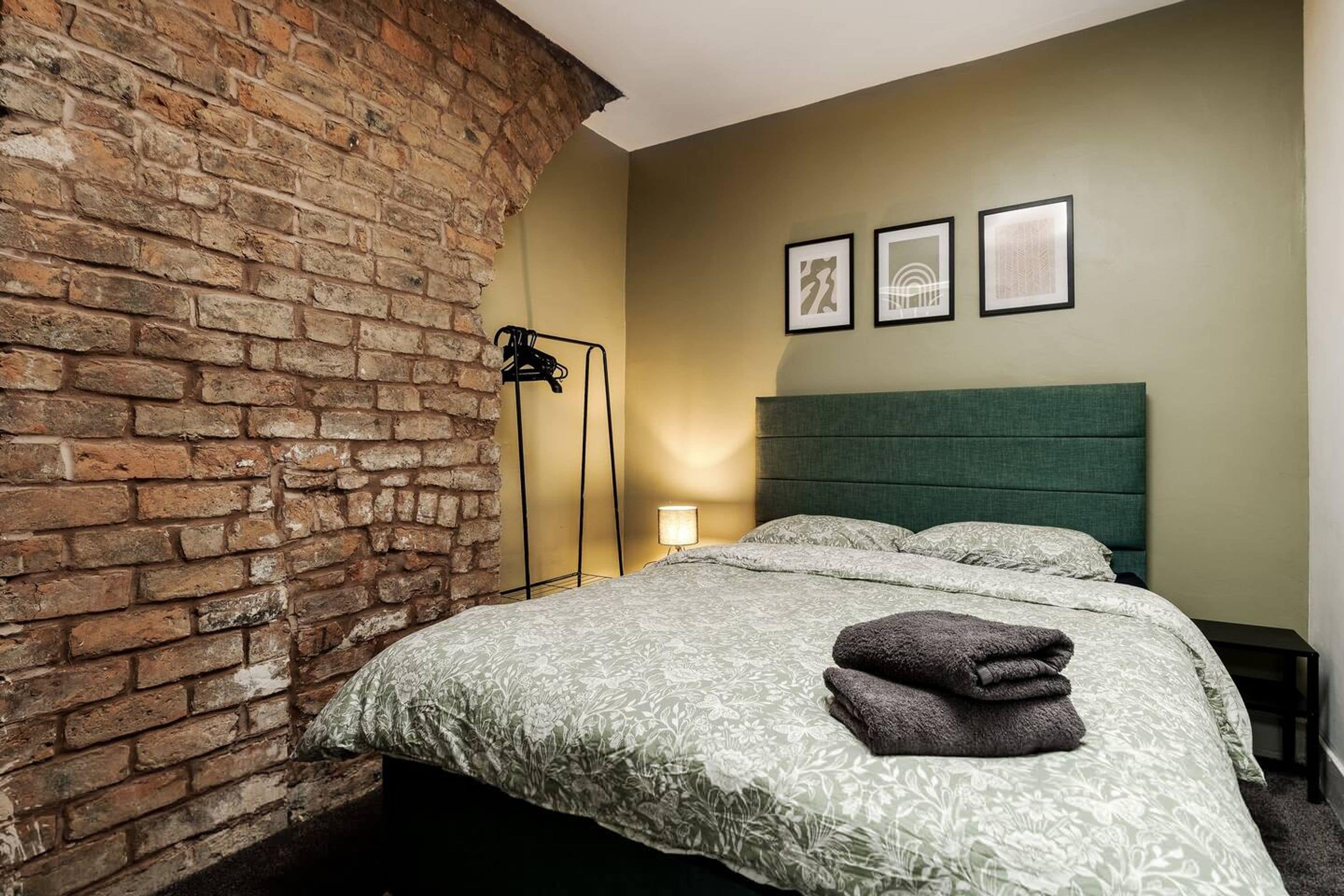 Een hotelkamer met een bakstenen muur, een bed met een groen hoofdeinde en drie ingelijste foto's aan de muur.