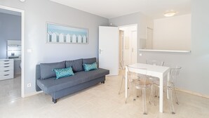 Ett modernt vardagsrum med en grå soffa, ett matbord med genomskinliga stolar, en stor spegel och en tavla på väggen.