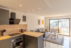 A modern kitchen with a built-in oven, a dining area with a round table, and a large window offering a view of the city. - 1 Bedroom Renovated Near Monaco (Beausoleil)