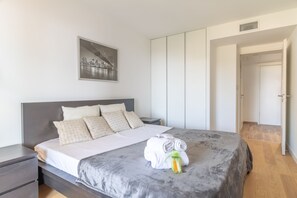 Ein ordentlich bezogenes Bett mit grauer Decke, einem Handtuch und einer Wasserflasche. Ein Nachttisch mit Lampe steht neben dem Bett, und ein gerahmtes Bild hängt an der Wand.