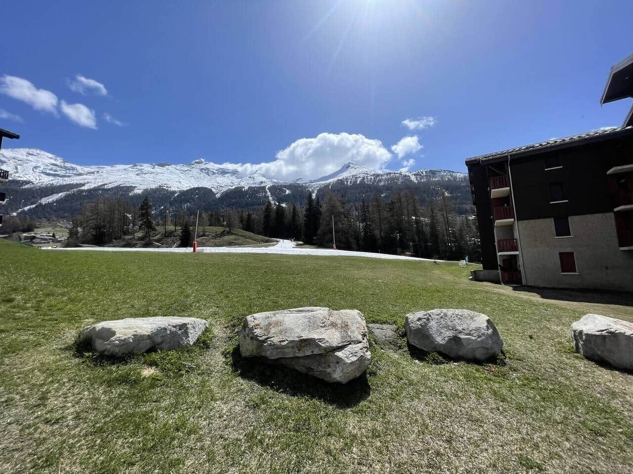 A grassy area with large rocks, a building with balconies, and snow-capped mountains in the background.