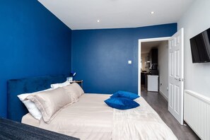 A bedroom with a bed, pillows, a nightstand, a TV, and a kitchenette in the background. - Relaxing Annexe With Garden Access Parking (Maidstone)