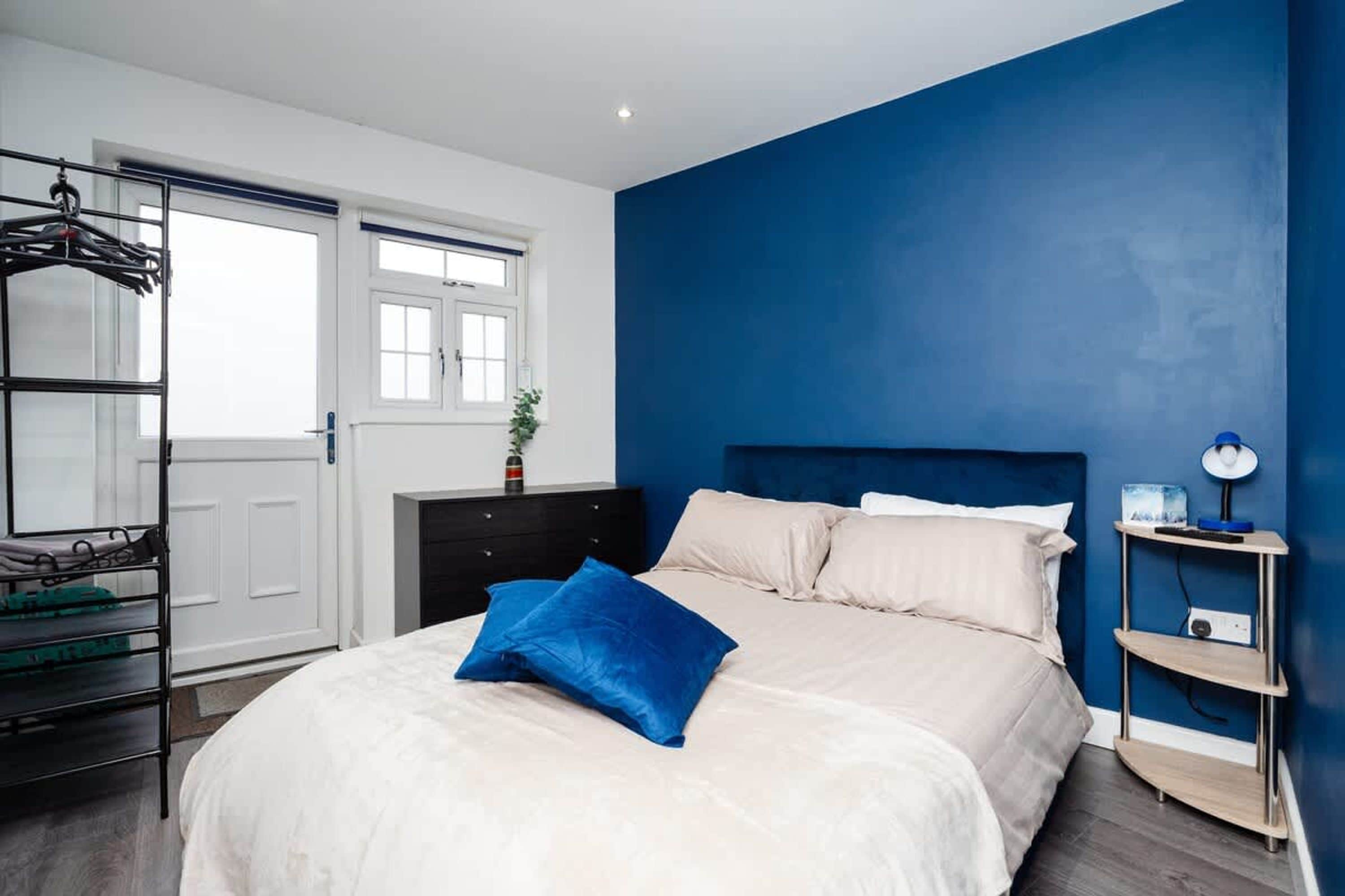 Un dormitorio con cama, pared azul, puerta blanca, cómoda y estante con lámpara.