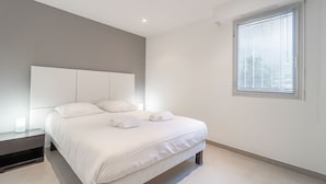 A modern bedroom with a large bed, a bedside table, a window with blinds, and a ceiling light. - Stunning Renovated 1-bedroom Breathtaking View (Beausoleil)