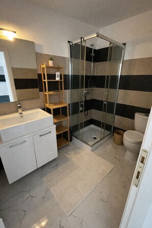 Ein Badezimmer mit einer Duschkabine, einem weißen Waschbecken, einem Holzregal und einer Toilette.