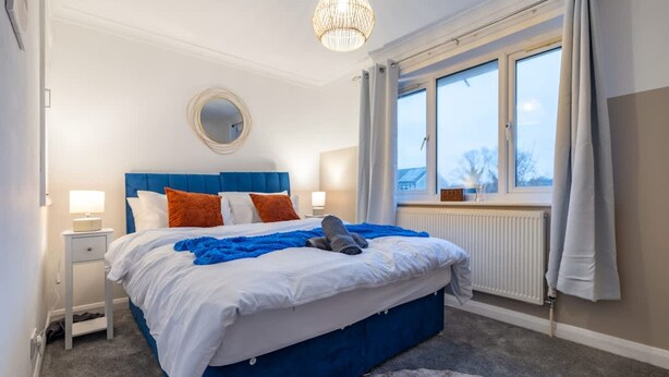 A bedroom with a blue headboard, white bedding, a round mirror, a bedside table with a lamp, a window with curtains, and a view of the outside. - Perfect Booking 2 Bed Home in Hitchin (Hitchin)