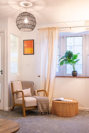 A room with a wicker chair, a small ottoman, a hanging wicker light fixture, a window with a curtain, and a potted plant.