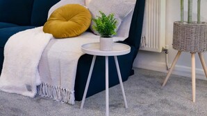 A living room with a dark blue sofa, a small white side table, a potted plant, and a radiator. - Perfect Booking 2 Bed Home in Hitchin (Hitchin)