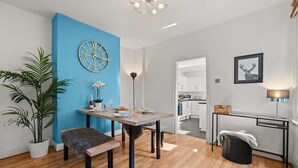 A modern dining area with a blue accent wall, a round wall clock, a dining table with chairs, a bench, a potted plant, and a desk with a lamp. - Big Family Home Free Parking Fast Wifi Garden (Kingston Upon Thames)